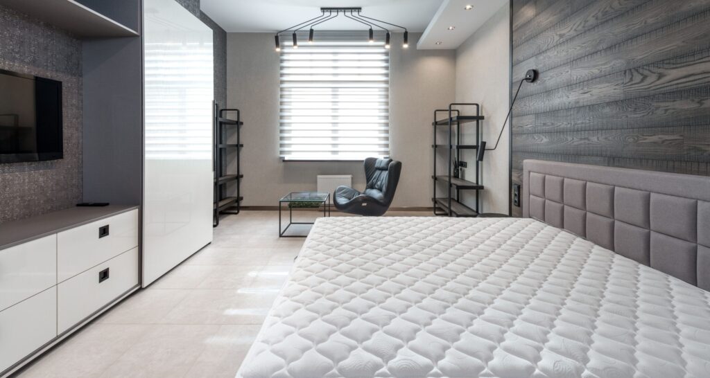 Bed with mattress placed against wall with modern TV and white cupboards in stylish bedroom with window and leather armchair